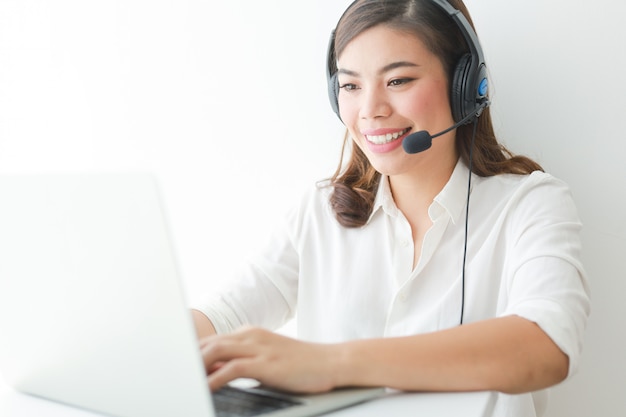 Azjatycka kobieta na białej koszula mówi z hełmofonem i używa laptopu, uśmiechu i szczęśliwego twarz operatora pojęcia