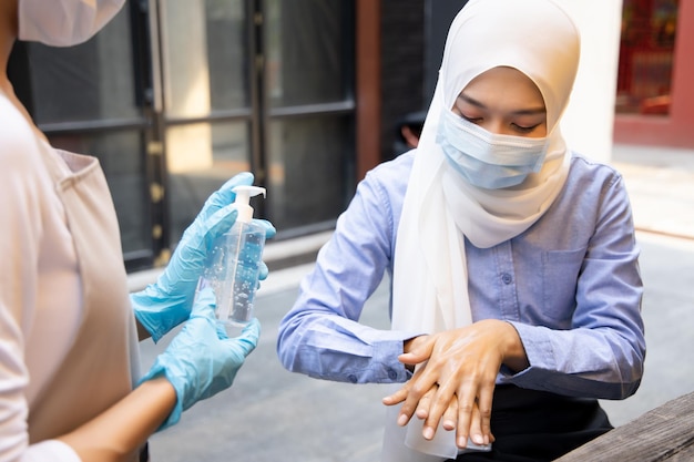 Zdjęcie azjatycka kobieta, pracownica lub właścicielka sklepu, nosząca gumową rękawiczkę podczas dozowania alkoholowego żelu do dezynfekcji rąk muzułmańskiej klientce, koncepcja nowej normalnej praktyki biznesowej z integracyjną różnorodnością ludzi