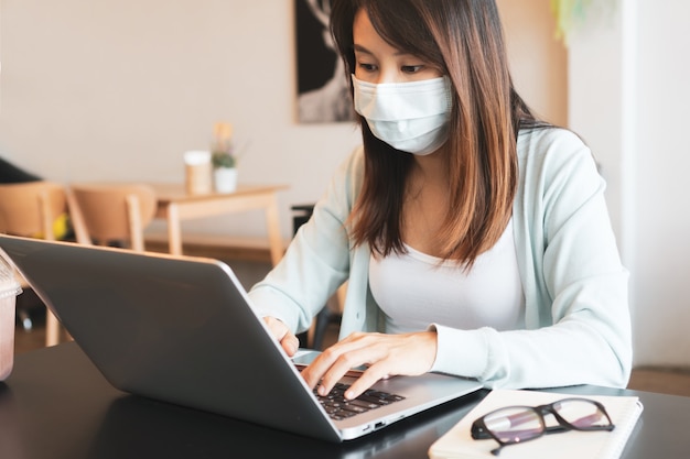 azjatycka kobieta pracująca na komputerze podczas noszenia maski medycznej w kawiarni podczas kryzysu związanego z koronawirusem