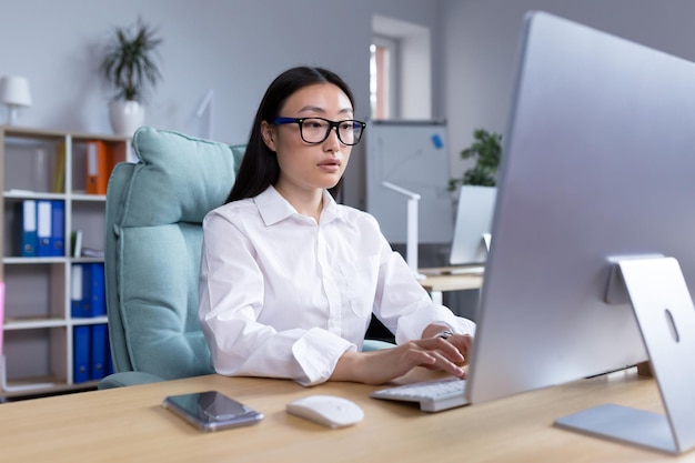 Azjatycka kobieta pracuje w nowoczesnym biurze przy komputerze, wpisując raport online