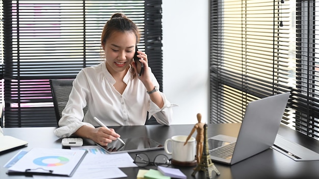 Azjatycka kobieta rozmawia przez telefon komórkowy i pracuje z laptopem w biurze