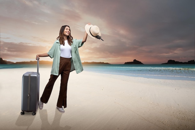 Azjatycka kobieta stoi na plaży w kapeluszu i walizce