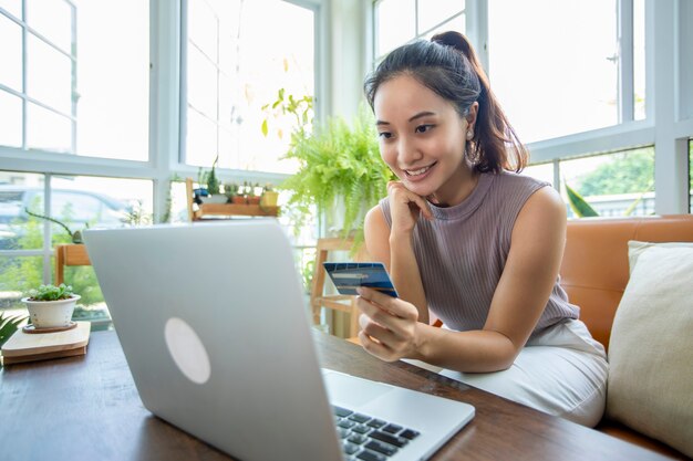 Azjatycka kobieta trzyma kartę kredytową i używa smartfona do zakupów online w domu.