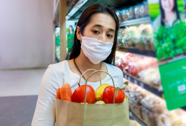 Azjatycka Kobieta Ubrana W Ochronną Maskę Na Twarz Trzyma Papierową Torbę Na Zakupy Z Owocami I Warzywami W Supermarkecie.