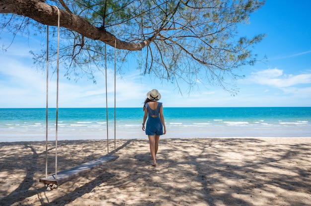 Azjatycka kobieta w kapeluszu spaceru na plaży i drewniana huśtawka zwisająca z drzewa w tropikalnym morzu