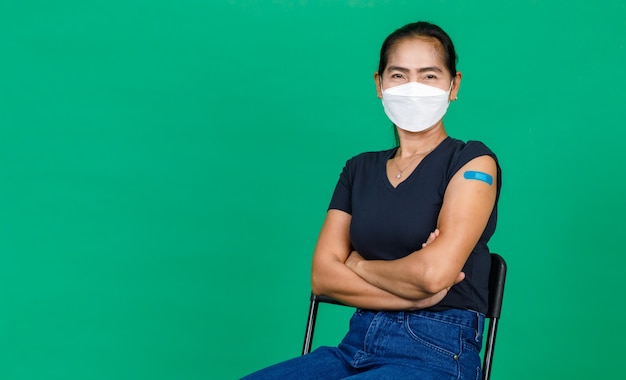 Azjatycka kobieta w średnim wieku nosząca maskę pokazującą ramię z bandażem pokazującym, że została zaszczepiona na wirusa Covid 19 na zielonym tle. Koncepcja szczepienia przeciwko Covid 19.