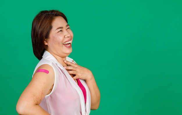 Azjatycka kobieta w średnim wieku śmiejąca się z ulgą, jej ramię z bandażem pokazuje, że została zaszczepiona na wirusa Covid 19 na zielonym tle. Koncepcja szczepienia przeciwko Covid 19.