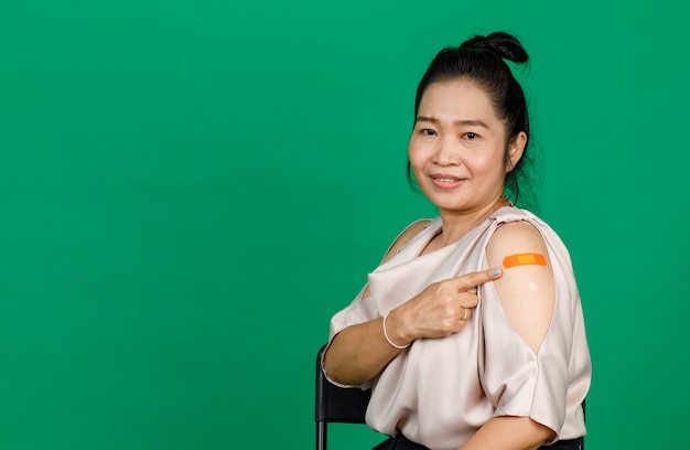 Azjatycka kobieta w średnim wieku uśmiechnięta i wskazująca na ramię z bandażem pokazującym, że została zaszczepiona na wirusa Covid 19 na zielonym tle. Koncepcja szczepienia przeciwko Covid 19.