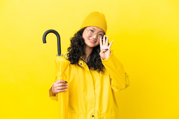 Azjatycka kobieta z płaszczem przeciwdeszczowym i parasolem na żółtym tle szczęśliwa i licząca cztery palcami