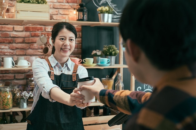 azjatycka koreańska kobieta barista w dżinsowym fartuchu podająca gorącą kawę w białym papierowym kubku na wynos klientowi płci męskiej. sklep z kawiarnią na wynos. uśmiechnięta młoda kelnerka z dwiema rękami serwująca napój na wynos