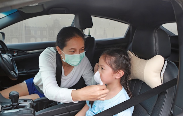 Azjatycka matka i noszenie higienicznej maski na twarz dla córki siedzącej w samochodzie podczas epidemii koronawirusa (covid-19)