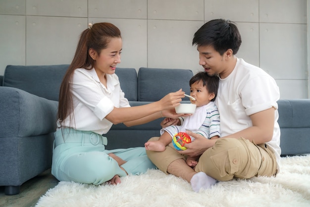 Azjatycka matka karmi swojego 6-miesięcznego chłopca stałym pokarmem z łyżeczką, a ojciec siedzi obok, aby dopingować syna do jedzenia w salonie w domu.
