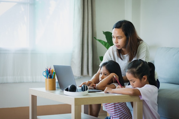 Azjatycka matka wspiera i pomaga dwóm córkom uczyć się online na laptopie w salonie. Rodzeństwo uczy się w domu podczas kwarantanny z powodu pandemii Covid 19.