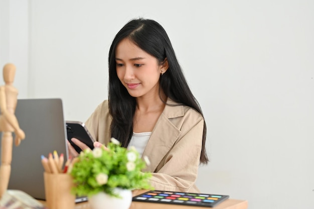 Azjatycka millenialsi businesswoman siedzi przy biurku i korzysta ze smartfona