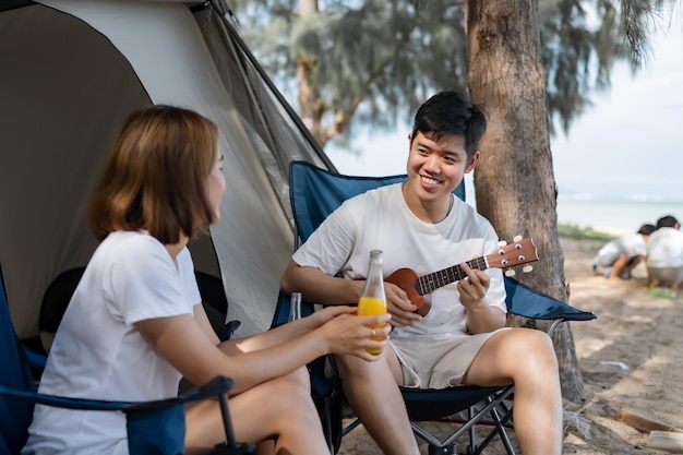 Azjatycka Miłość Para Kemping Na Plaży Latem. Mężczyzna Grający Na Ukulele I Razem Z Kobietą Siedzącą Na Zewnątrz Namiotu.