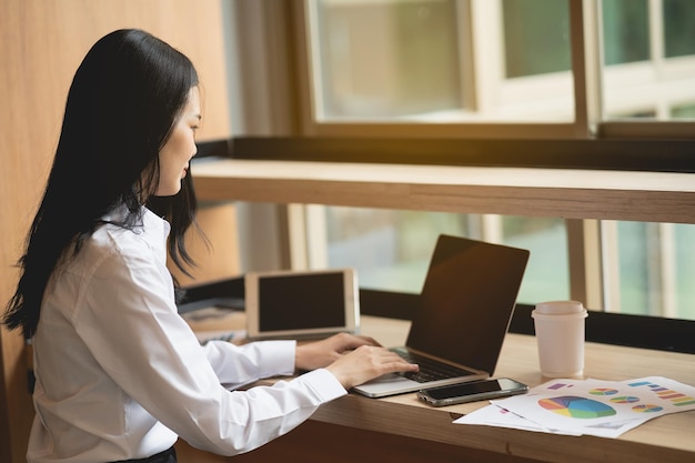 Azjatycka młoda kobieta w eleganckiej białej koszuli nosić pracuje na laptopie, siedząc w kreatywnym biurze lub ca