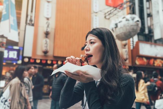 Azjatycka młoda modelka jedzenie knedle z ośmiornicą na tętniącej życiem okolicy dotonbori osaka w japonii. miejscowa dama je takoyaki uliczne jedzenie z wesołą uśmiechniętą twarzą. ogromny balon z rybą puffer z tyłu