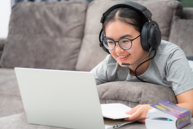Azjatycka młoda studentka ze słuchawkami leżąca na kanapie uśmiech patrząc na laptopa online nauka ze szkoły dziewczyna jest szczęśliwa na uczelnię internet studium na odległość online praca na komputerze z domu