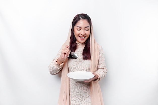 Azjatycka muzułmanka pości i jest głodna, trzymając sztućce, patrząc w bok, myśląc o tym, co zjeść