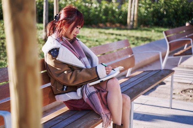 Azjatycka nastolatka siedzi na ławce w kampusie uczelni i pisze w swoim notatniku