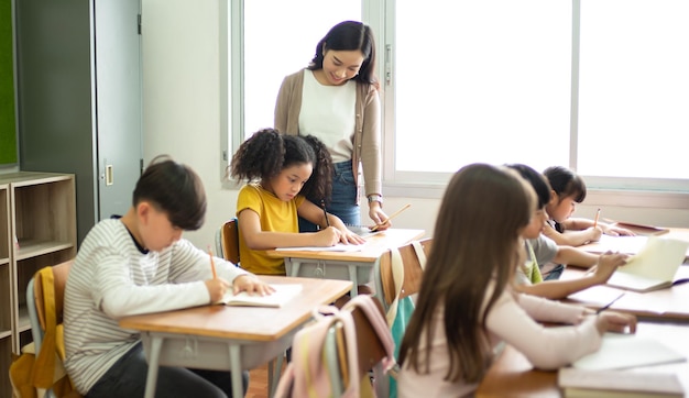 Azjatycka nauczycielka pomagająca uczennicy podczas pisania w notatniku na lekcjach w szkole podstawowej Uczniowie podczas egzaminu w klasie Koncepcja wiedzy o edukacji