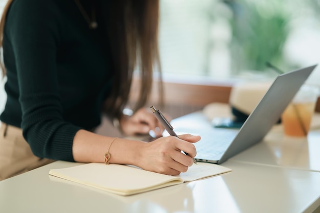 Azjatycka niezależna kobieta uśmiecha się za pomocą pióra robienia notatek w swoim notatniku i pracuje na laptopie na stole w domu Kobieta przedsiębiorca pracująca dla swojej firmy w kawiarni Koncepcja biznesowa w kawiarni