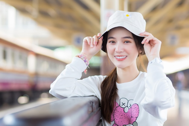 Azjatycka piękna dziewczyna w białej koszuli z długimi rękawami i kapeluszu siedzi szczęśliwa buźka na stacji kolejowej