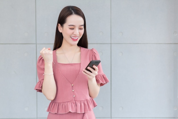 Azjatycka piękna kobieta w różowej sukience uśmiecha się szczęśliwie, trzymając smartfon.