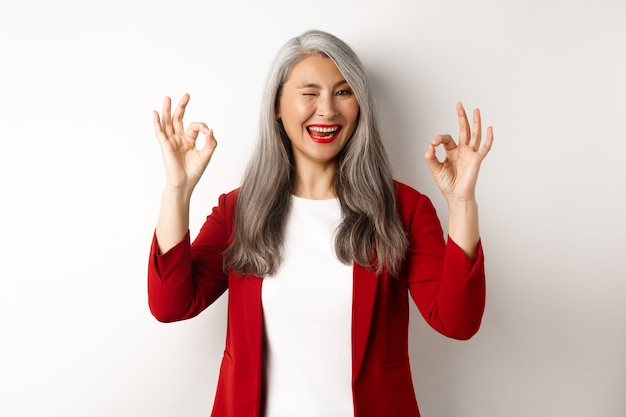 Azjatycka Profesjonalna Bizneswoman Pokazująca Znaki Ok I Mrugająca, Uśmiechnięta Zadowolona, Zapewniająca Lub Polecająca Coś, Stojąca Na Białym Tle