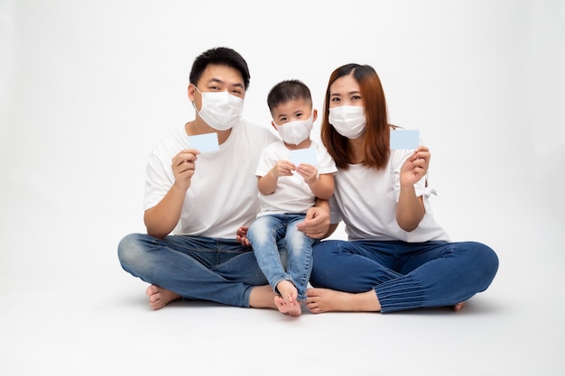 Azjatycka rodzina jest ubranym ochronną maskę medyczną dla zapobiegania wirusowi Covid-19 i trzyma ubezpieczeniową kartę opieki odizolowywającą na biel ścianie. Koncepcja ochrony rodziny i ubezpieczenia karty medycznej