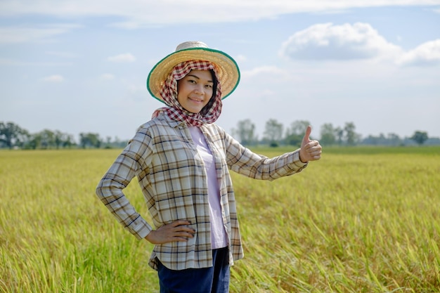 Azjatycka rolniczka ubrana w pasiastą koszulę w kapeluszu stojąca i pozująca kciuk do góry z uśmiechniętą twarzą na polu ryżowym