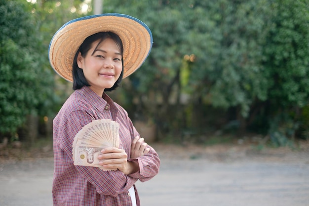 Azjatycka rolniczka z ramieniem krzyżowym i trzymająca pieniądze z banknotów tajlandzkich na farmie