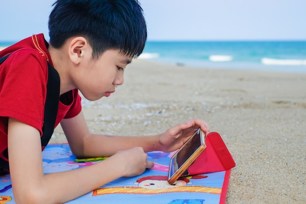 Azjatycka śliczna chłopiec używa pastylkę dla sztuki gry na plaży