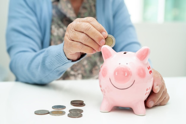 Azjatycka starsza kobieta wkłada monetę do różowej skrzynki na oszczędności i ubezpieczenia ubóstwo problemy finansowe na emeryturze