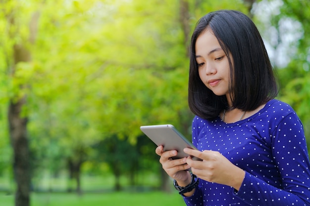 Azjatycka studencka dziewczyna używa cyfrową pastylkę w parku w pogodnym letnim dniu