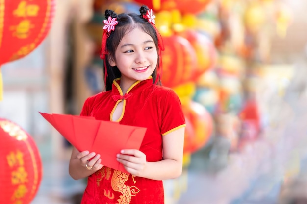 Azjatycka Szczęśliwa dziewczynka ubrana w czerwoną tradycyjną chińską dekorację cheongsam trzymająca w ręku czerwone koperty i latarnie z chińskim tekstem Błogosławieństwa to błogosławieństwo dla Chińczyków