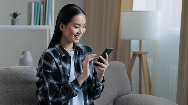 Azjatycka szczęśliwa uśmiechnięta dziewczyna kobieta siedzi w domu na kanapie patrząc na telefon komórkowy na czacie przeglądanie wifi