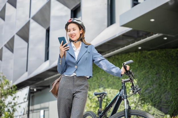 Azjatycka uśmiechnięta młoda kobieta w kasku trzyma mobilną inteligentną rozmowę telefoniczną z biznesem podczas pracy biurowej na ulicy z rowerem, przyjazna dla środowiska, biznesowa kobieta dojeżdżająca do pracy rano na zewnątrz