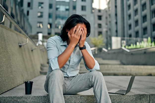 Azjatycki Biznesmen W Mieście. Zdenerwowany Młody Człowiek Siedzący Na Schodach W Pobliżu Centrum Biznesowego Z Laptopem