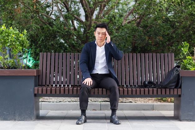 Azjatycki biznesmen zmęczony po pracy, siedzący na ławce ze zmęczenia i bólem głowy, ma stres i ataki paniki