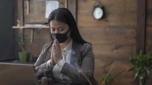 Azjatycki bizneswoman ono Modli się W biurze Stawia Jej dłonie Wraz Z Zamkniętymi oczami Podczas gdy Siedzący Przy stołem, pandemiczny pojęcie