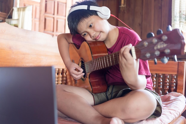 Azjatycki chłopiec noszący muzykę online na słuchawkach na swoim laptopie, używający twórców dzieciaka na gitarze akustycznej