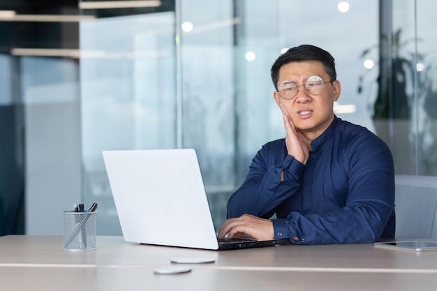 Azjatycki chory w biurze biznesmen ma ból zęba siedzącego w biurze wewnątrz budynku, w którym pracuje