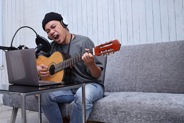 Azjatycki facet grający na gitarze, nagrywający podcast audio w studio za pomocą mikrofonu i laptopa. Ludzie