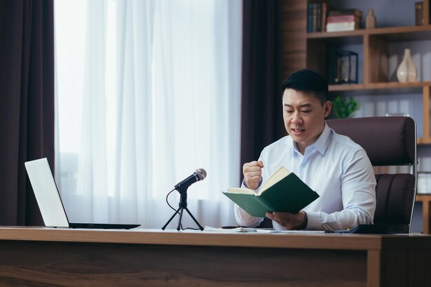 Azjatycki kaznodzieja czyta Biblię w Internecie nagrywa podcast siedzi przy stole i patrzy w kamerę używa mikrofonu trzyma rękę w sercu audycja radiowa online
