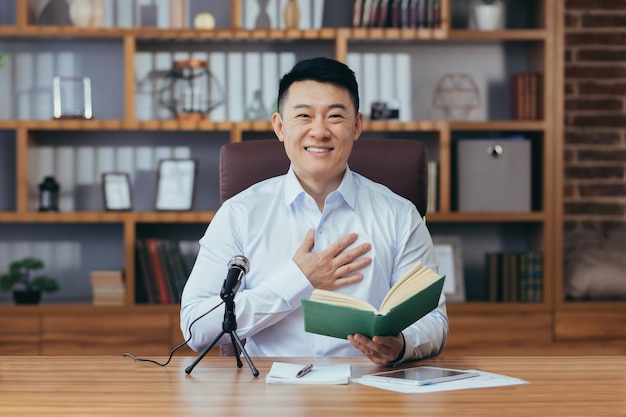 Azjatycki kaznodzieja czyta Biblię w Internecie nagrywa podcast siedzi przy stole i patrzy w kamerę używa mikrofonu trzyma rękę w sercu audycja radiowa online