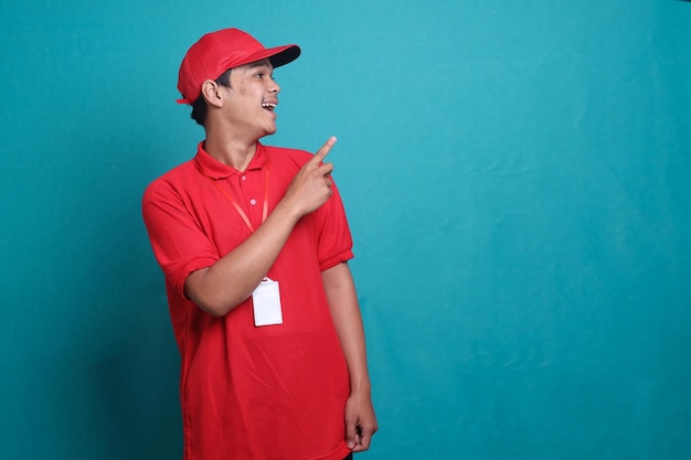 Azjatycki kurier w czerwonej koszulce i czapce uśmiechający się, wskazując palcem na puste miejsce. Ekspresowa przesyłka pocztowa