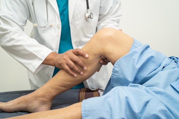 Azjatycki lekarz fizjoterapeuta badający masowanie i leczenie kolana i nogi starszego pacjenta w przychodni ortopedycznej pielęgniarki szpitalnej