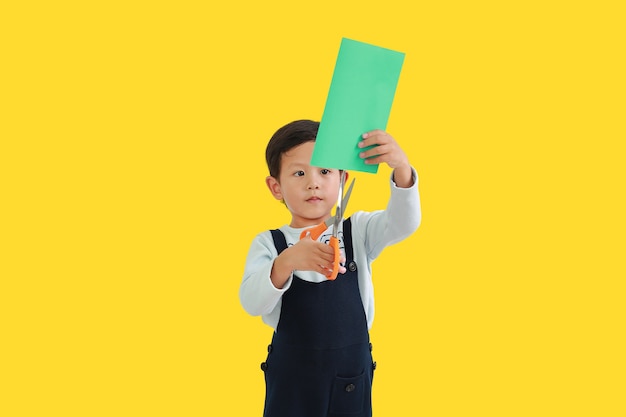 Azjatycki mały chłopiec lubi pracę rzemieślniczą nożyczkami wyciąć kolorowy papier na białym tle na żółtym tle. Obraz ze ścieżką przycinającą.