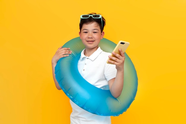 azjatycki mały chłopiec w wieku dziesięciu lat z nadmuchiwanym pierścieniem do pływania używa smartfona na żółtym tle